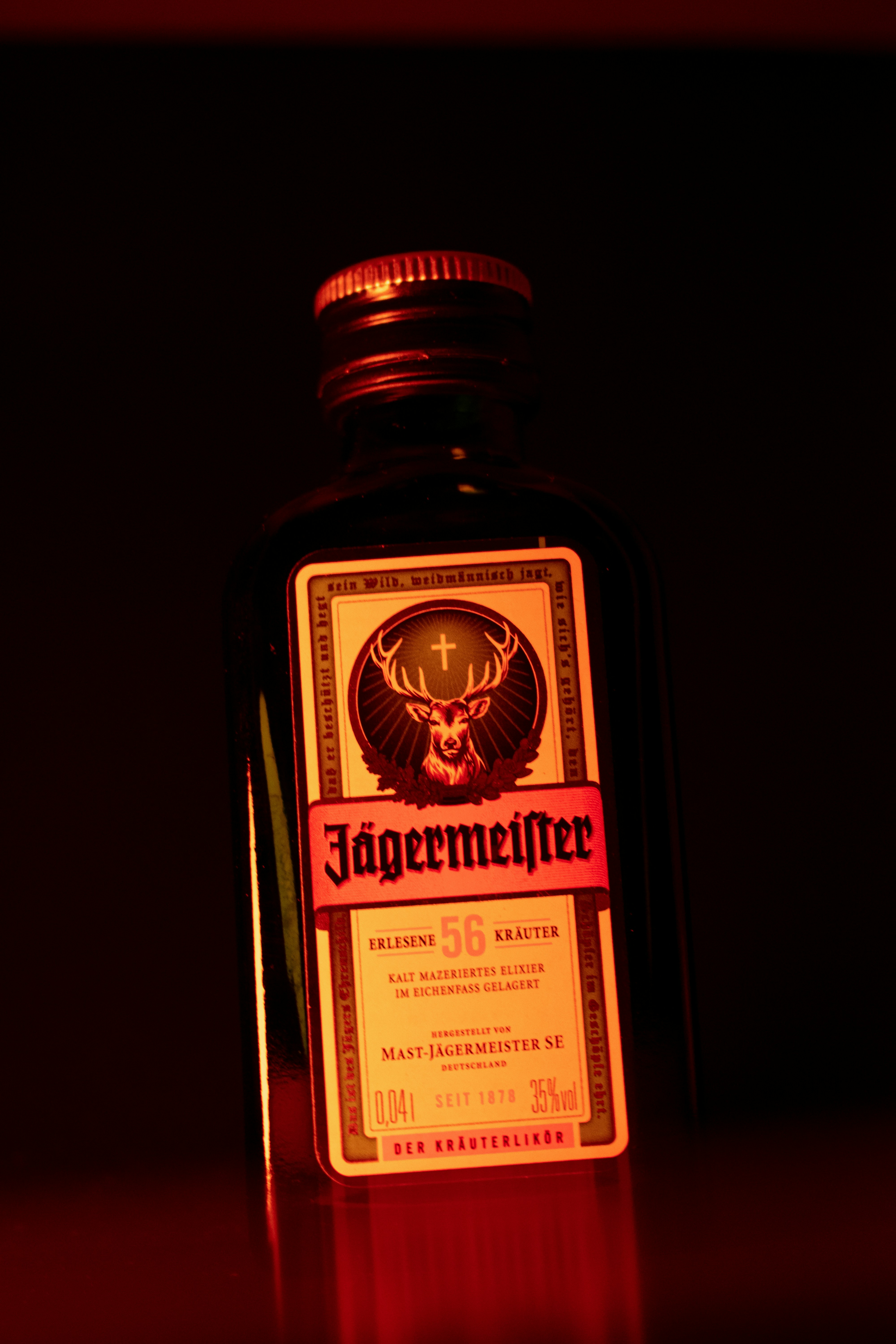 A bottle of Jägermeister liqueur, its label adorned with the brand's logo in elegant blackletter font, stands against a dark background illuminated by red lighting.