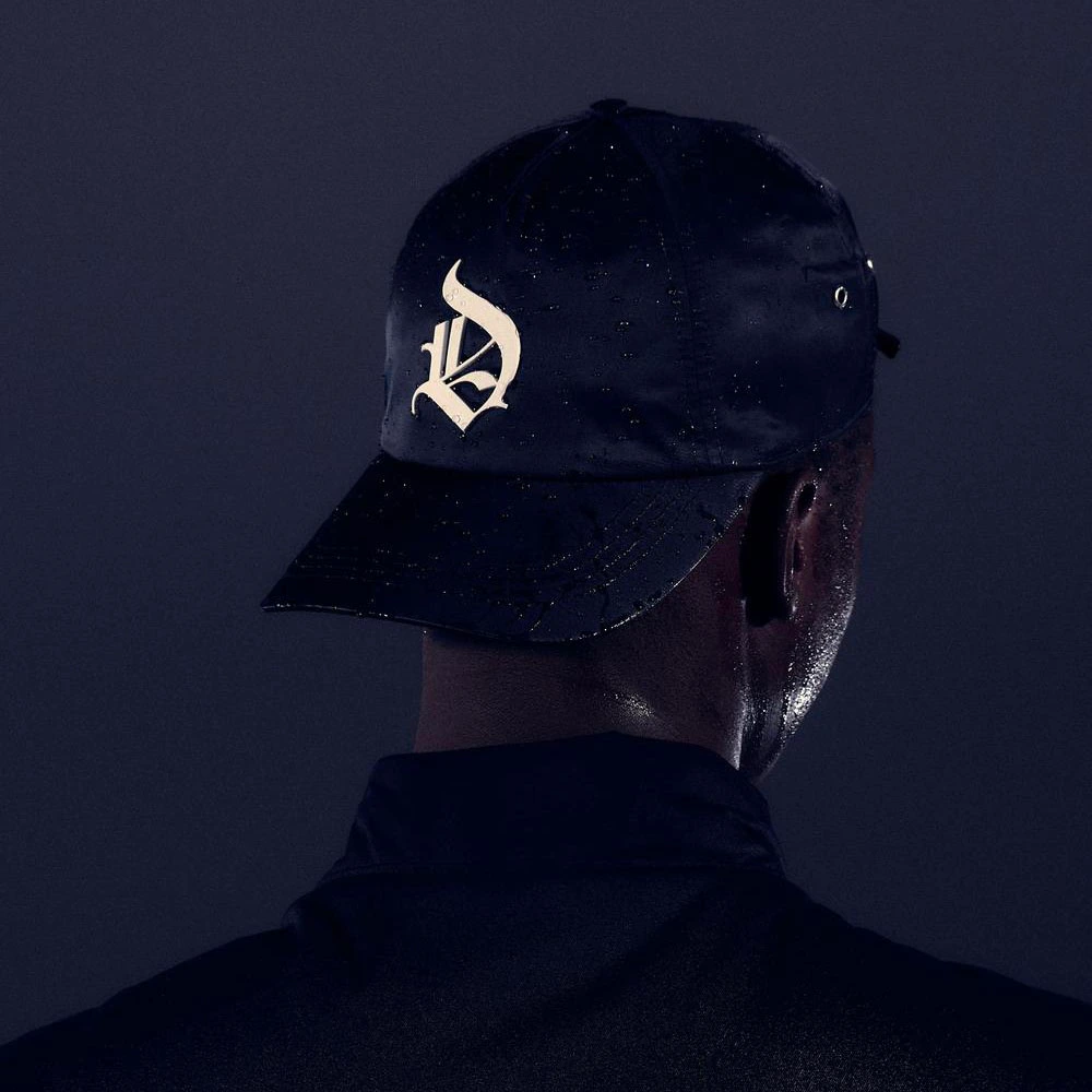 A person is shown from behind wearing a black cap with a white stylized 'D' logo and a dark jacket against a dark background